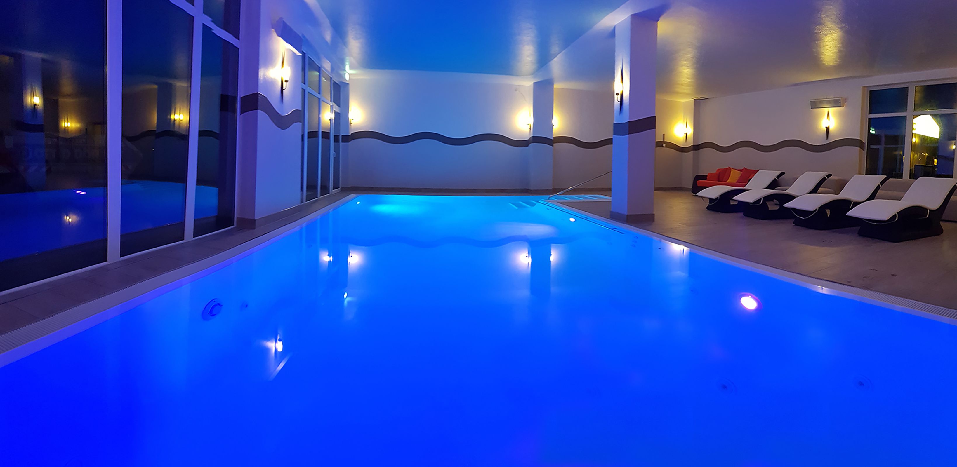 Una piscina con iluminación azul, tumbonas y apliques en la pared.