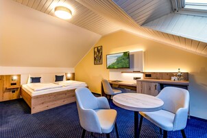 Quarto de hotel com cama, duas cadeiras, uma mesinha, escrivaninha e televisão.