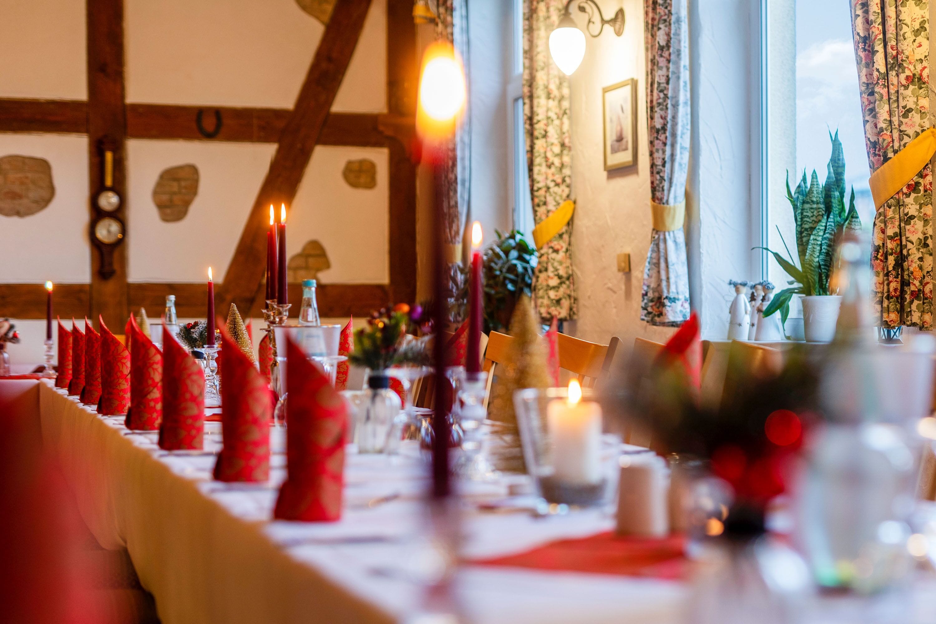 A dining table set with red napkins, candles, and glassware, in a room with exposed wooden beams and a window with curtains.