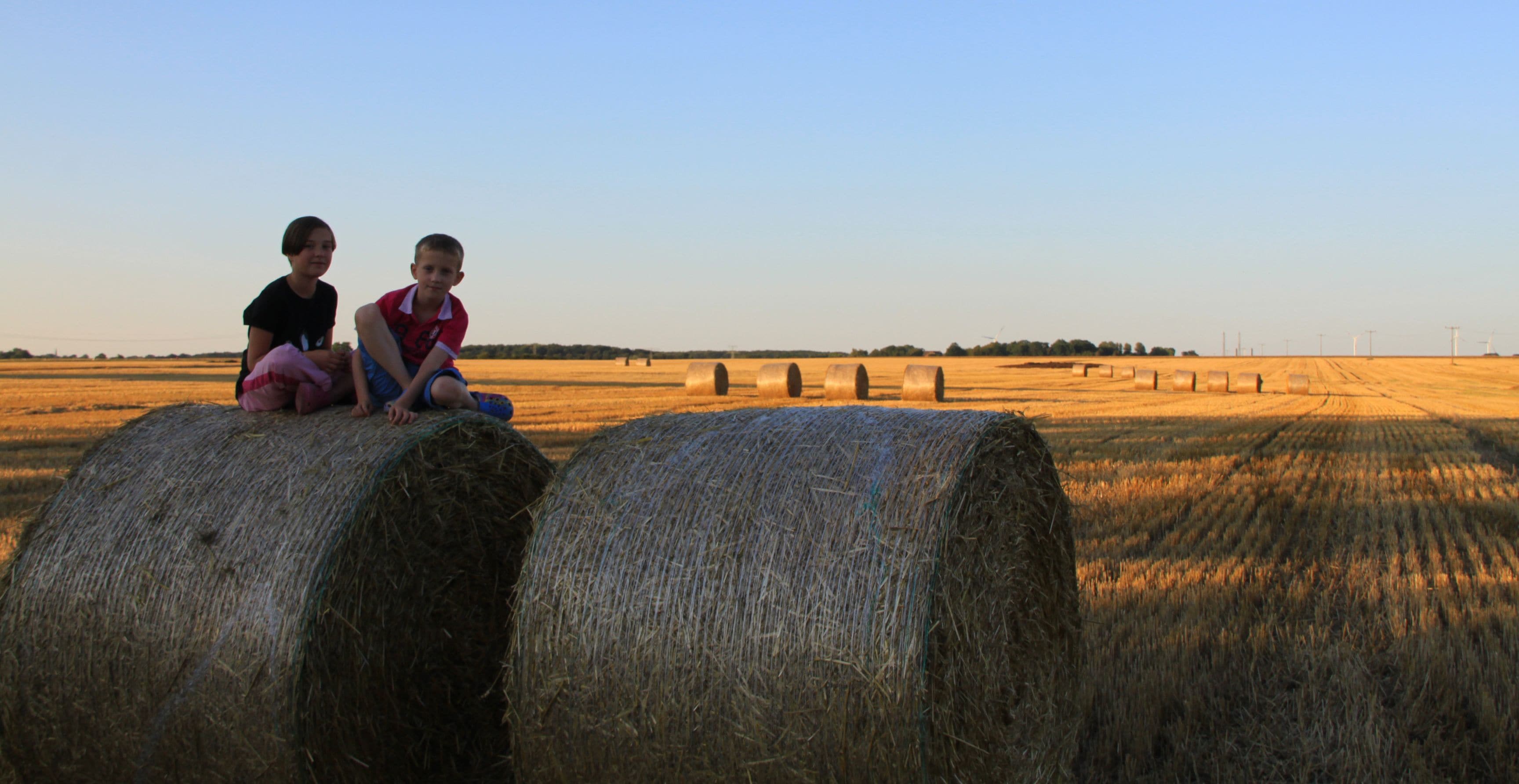 Two individuals sitting on hay bales in a field with hay bales and wind turbines in the background.