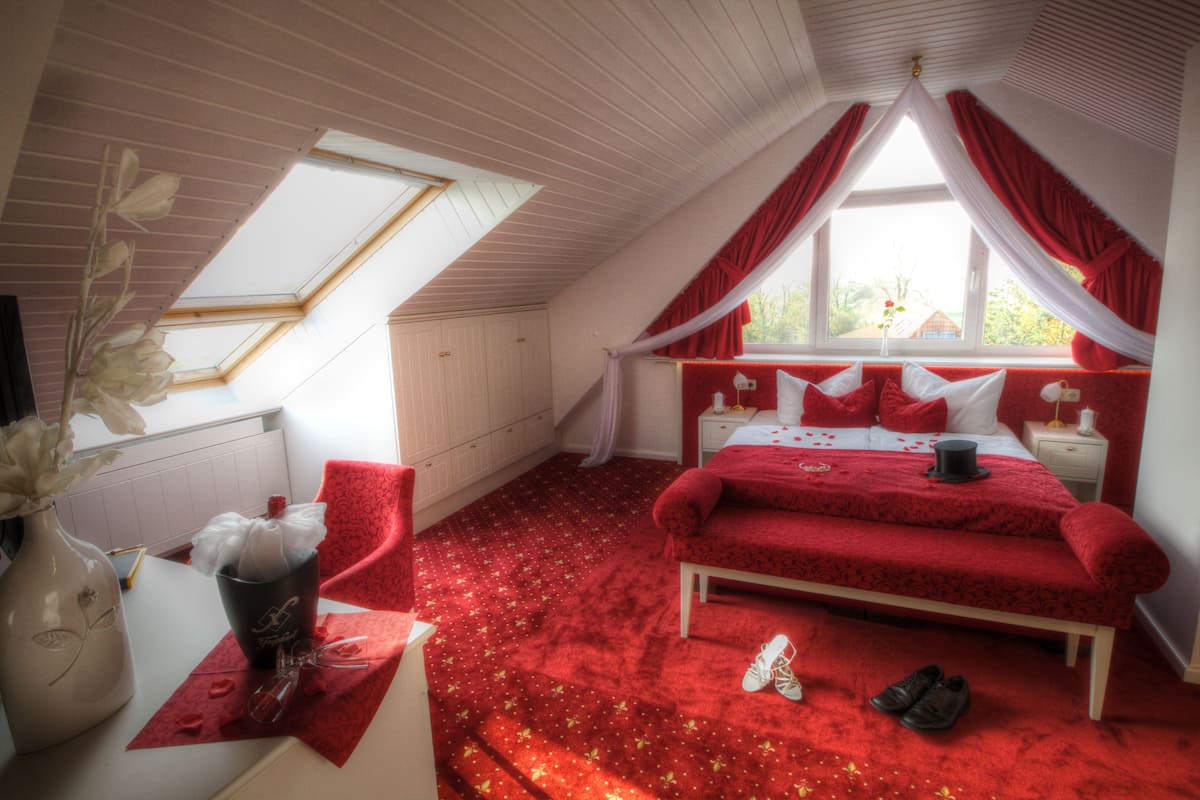 A bedroom with a red carpet, a bed with red bedding, a white dresser, a vase with flowers, and a window with red curtains.