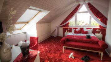 A bedroom with a sloped ceiling, a bed with red bedding, a nightstand, a chair, and a window with curtains.