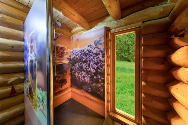 Interior de una cabaña de troncos con techo de madera, zona de ducha y un amplio ventanal con vistas a un área cubierta de césped.