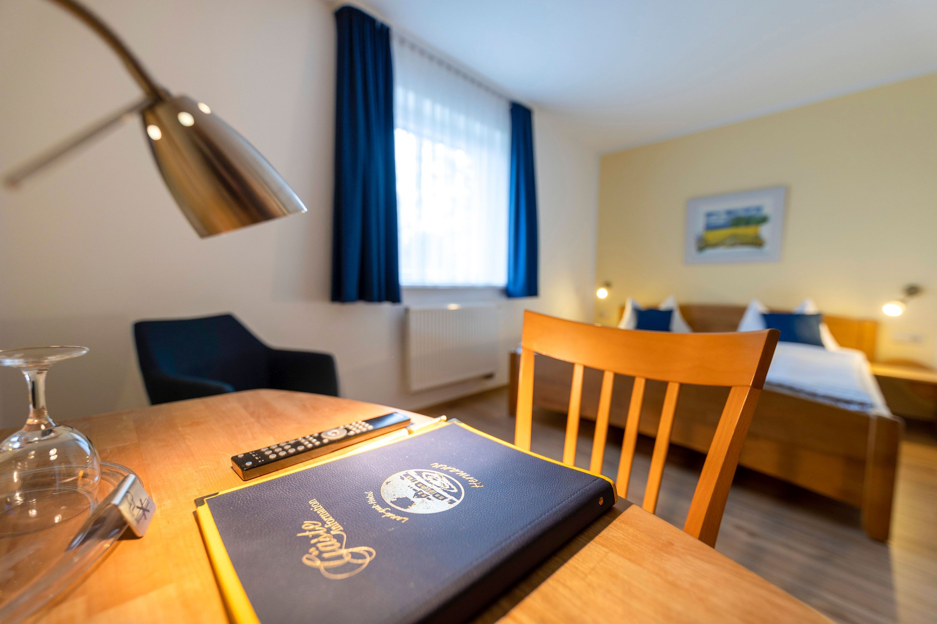 Ein Hotelzimmer mit einem Holztisch, einem Stuhl, einem Bett, einem Fenster mit Vorhängen und einer Lampe.