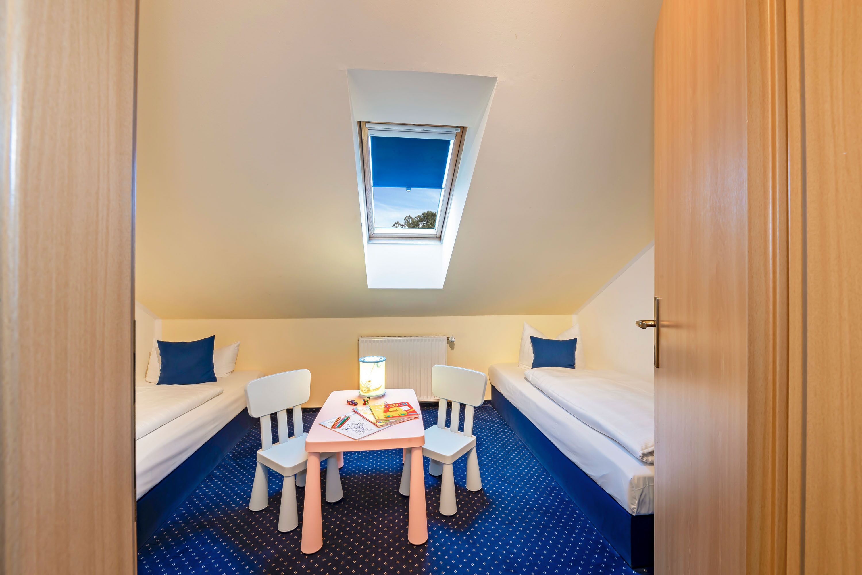 A compact room with a skylight, two single beds, a small table with a lamp, and a blue patterned carpet.