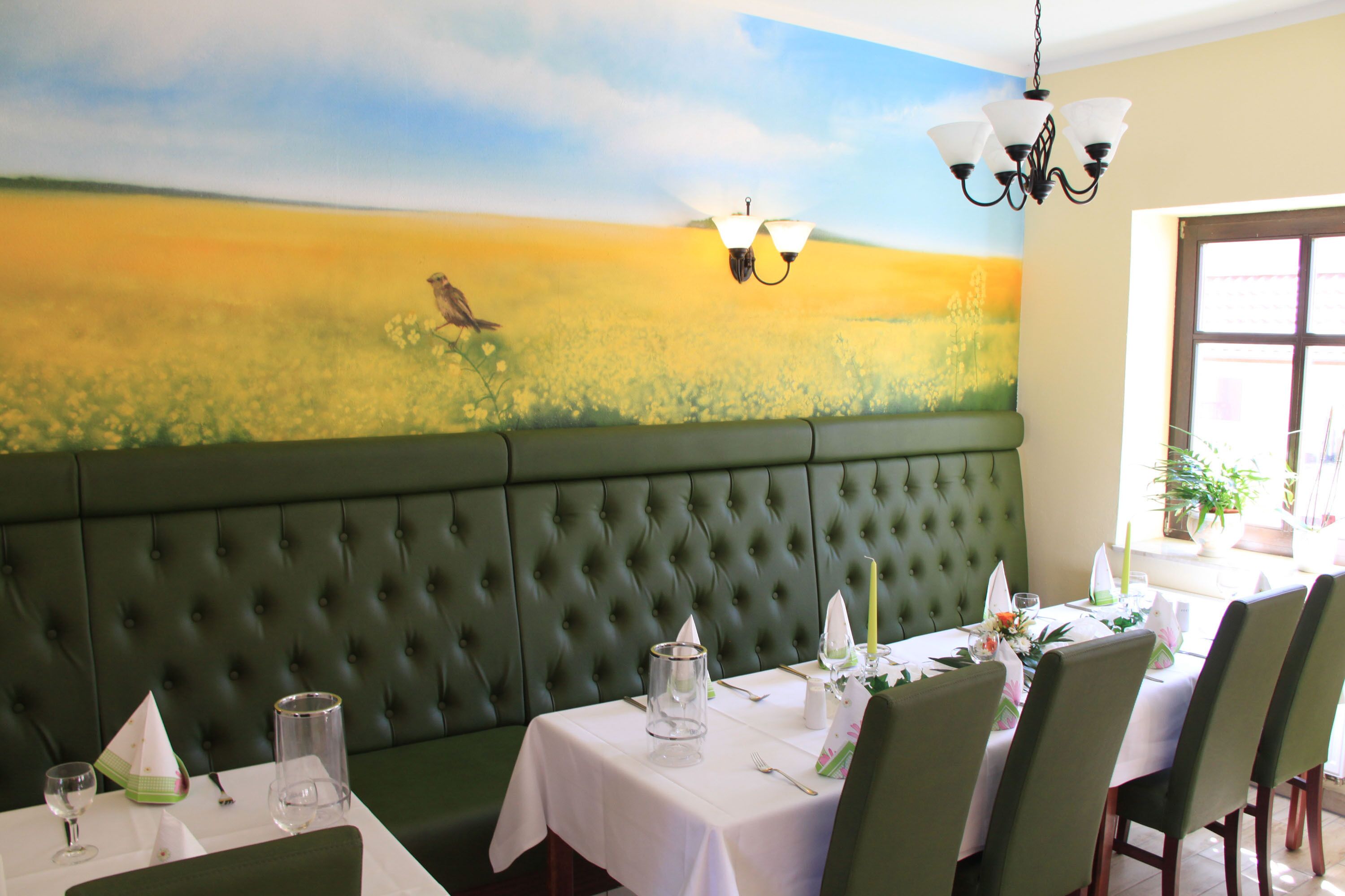 A dining area with a green upholstered booth, a table set with white tablecloths, and a mural of a field with a bird.