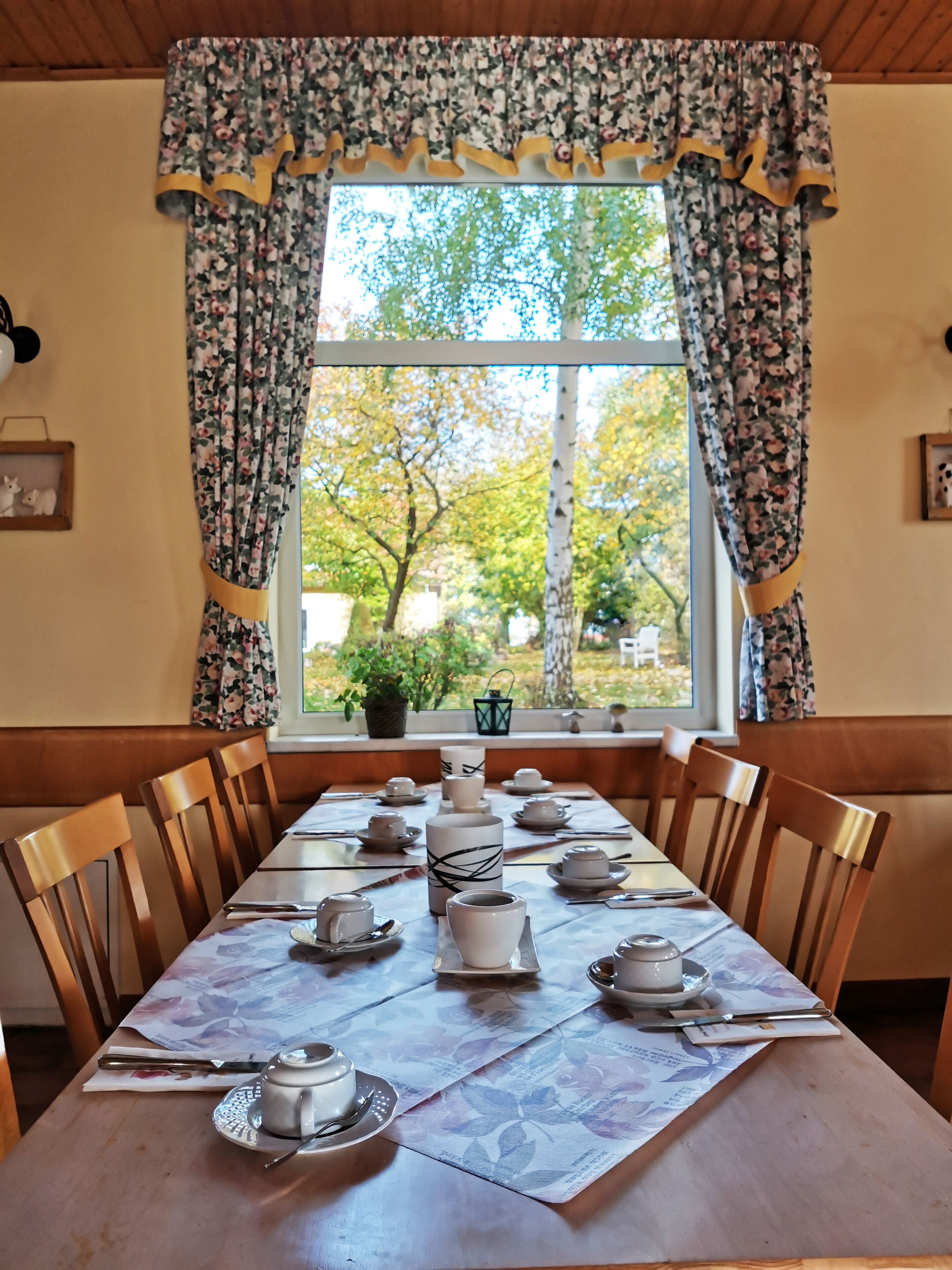 A dining table set for four with floral-patterned tablecloths and a window with floral curtains.
