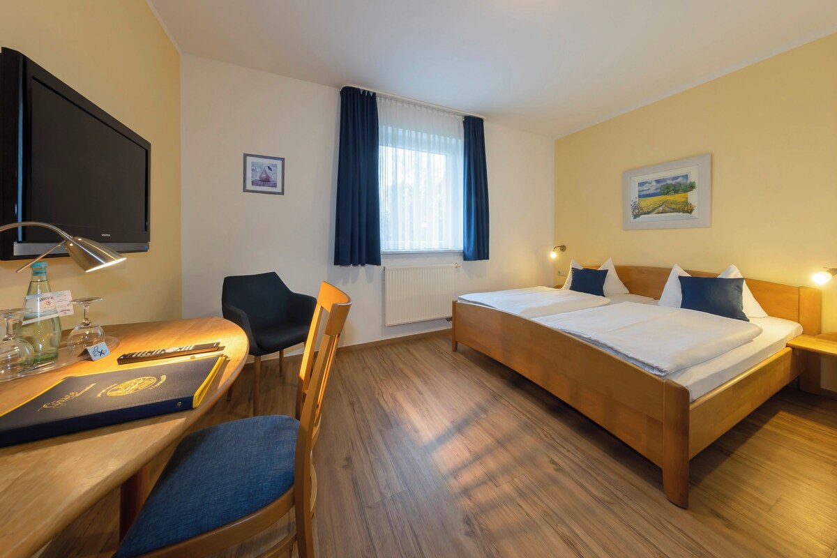 Una habitación de hotel con cama, escritorio con silla, televisor y ventana con cortinas.