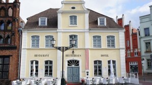 A building with a restaurant and hotel on the ground floor, featuring a central entrance with a blue door, white-framed windows, and outdoor seating with white chairs and tables. - Hotel und ReuterHaus (Wismar)