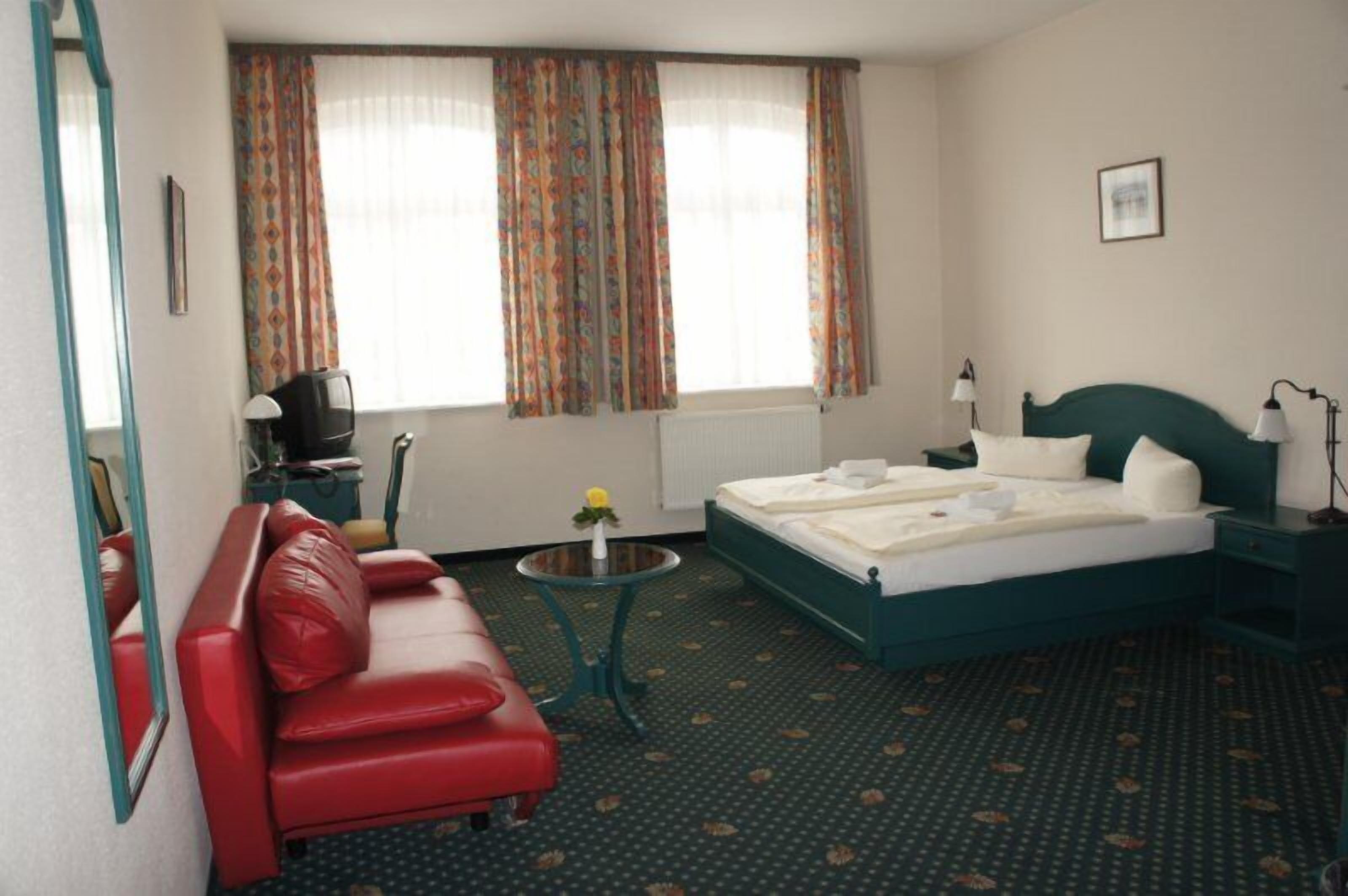 Ein Hotelzimmer mit einem Bett, einem roten Sofa, einem kleinen Tisch mit einer Vase und einem Fenster mit Vorhängen.