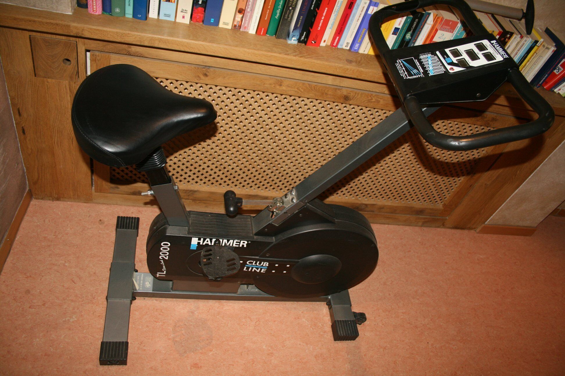 Een stilstaande hometrainer met een zwart zadel en handvatten, op een houten vloer met een geperforeerd houten paneel op de achtergrond.
