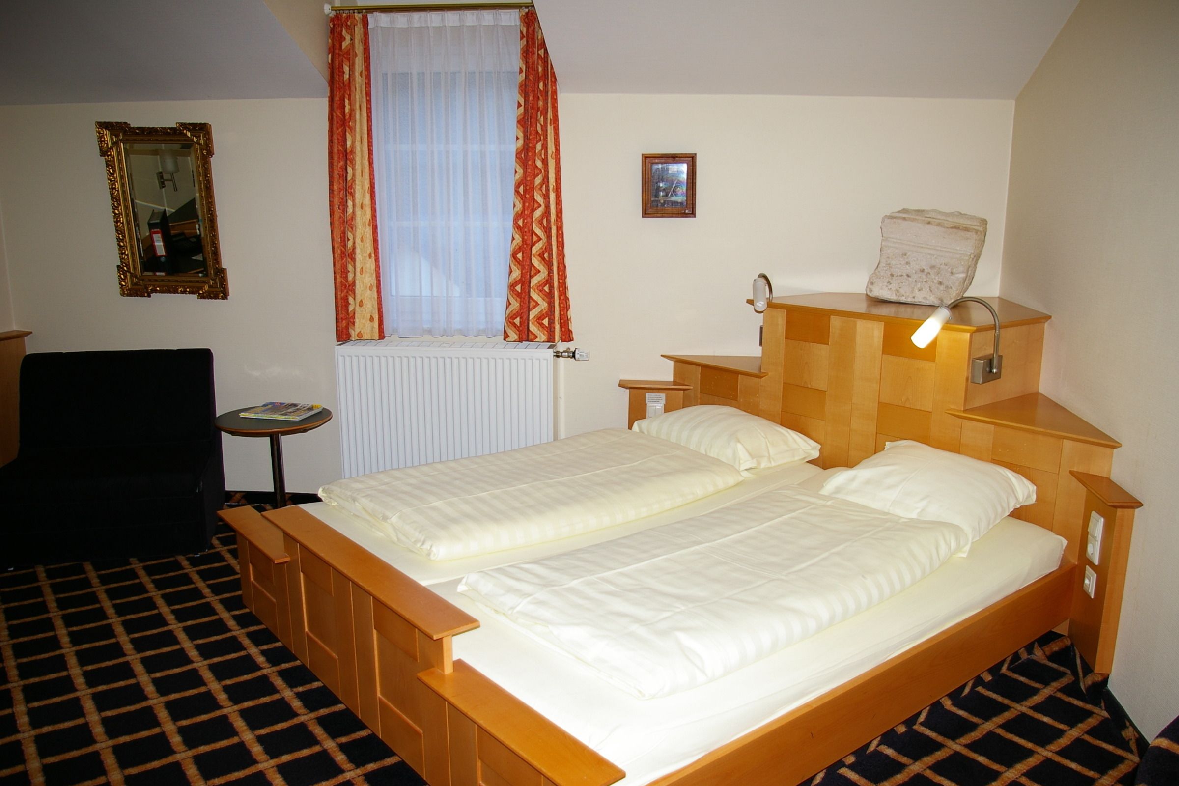 Habitación de hotel con cama doble, mesita de noche pequeña, una silla, ventana con cortinas, radiador y lámpara fijada a la pared.