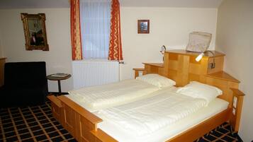 Habitación de hotel con cama doble, mesita de noche pequeña, una silla, ventana con cortinas, radiador y lámpara fijada a la pared.
