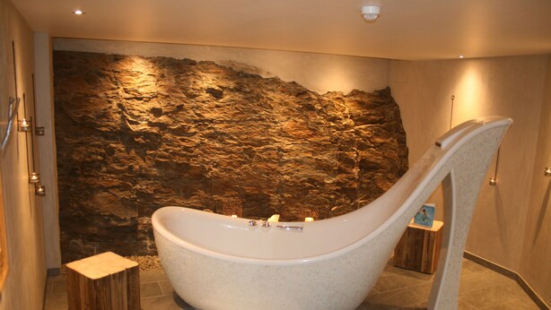 Een moderne badkamer met een groot bad, een houten kruk en een wand van gestructureerd steen.