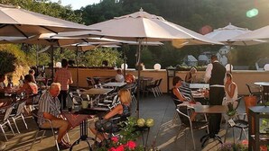 Een terras buiten met tafels en stoelen onder parasols, omringd door groen.
