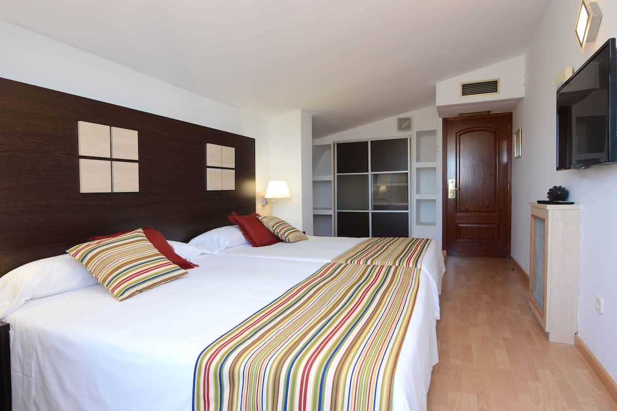 A hotel room with two beds, a wooden headboard, a TV mounted on the wall, and a hallway leading to a door.