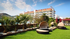 Un hotel con techo rojo, varios balcones y una zona de piscina con sillas de descanso y sombrillas.
