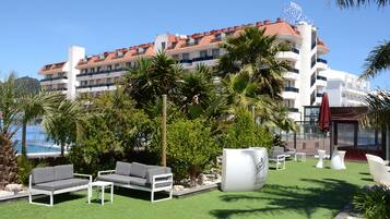 A hotel with a red-tiled roof, multiple balconies, and a pool area with artificial grass and outdoor furniture.