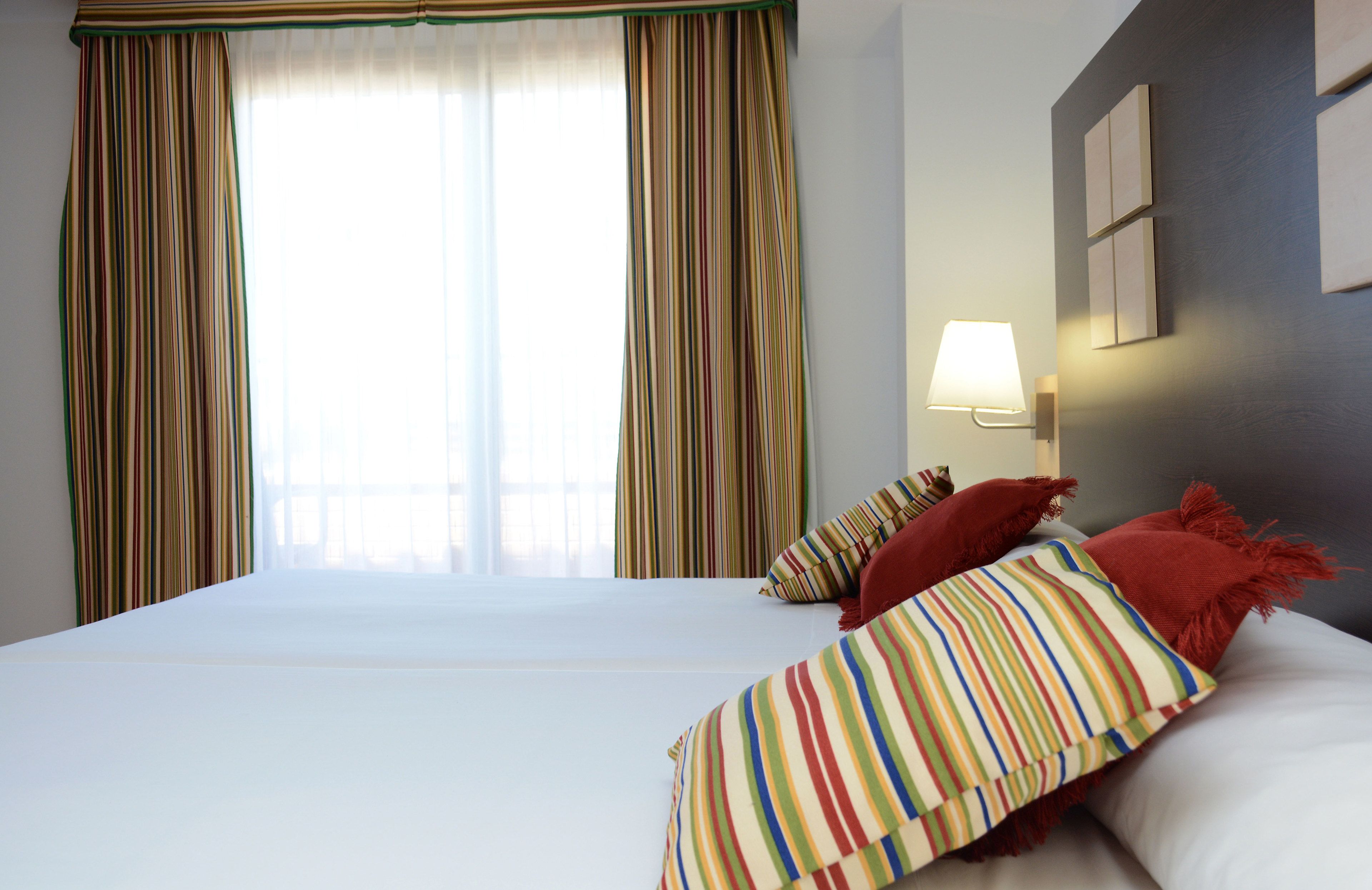 Habitación de hotel con una cama grande, cortinas a rayas, una lámpara de pared y vistas al exterior.