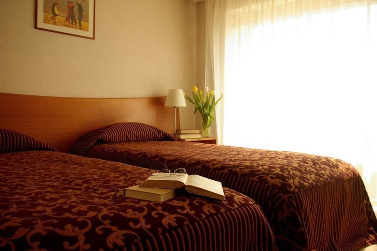 Una habitación de hotel con dos camas, una mesita de noche con una lámpara, un florero con flores y un cuadro en la pared.