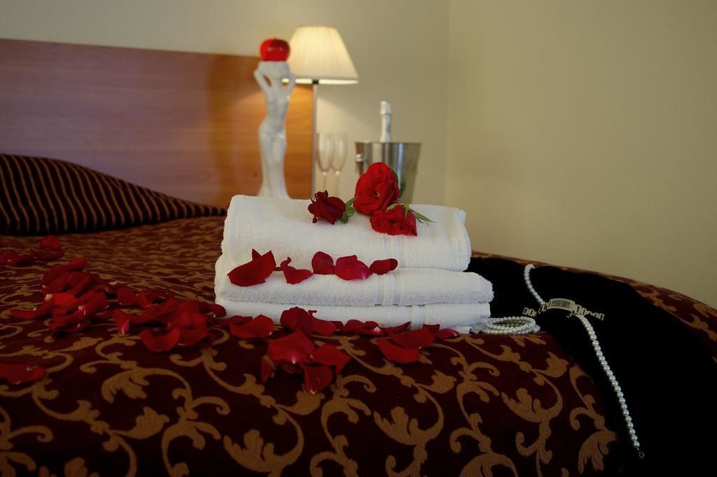 A neatly made bed with a white towel, a red rose, and rose petals. There is a white lamp and a glass of water on a side table.