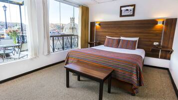 Une chambre d’hôtel avec un grand lit, une tête de lit en bois, une petite table de chevet, un banc et une vue sur la ville par une grande fenêtre.