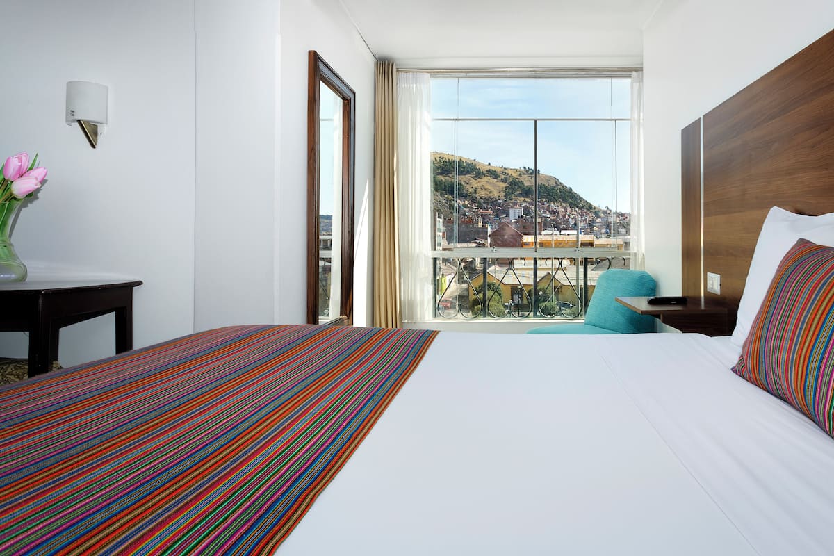 Une chambre d’hôtel avec un grand lit, une vue sur la ville, une tête de lit en bois et une petite table sur laquelle est posé un vase de fleurs.