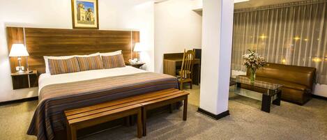 Een hotelkamer met een groot bed, een houten bankje, een glazen bijzettafeltje, een bank en een bureau met een lamp.