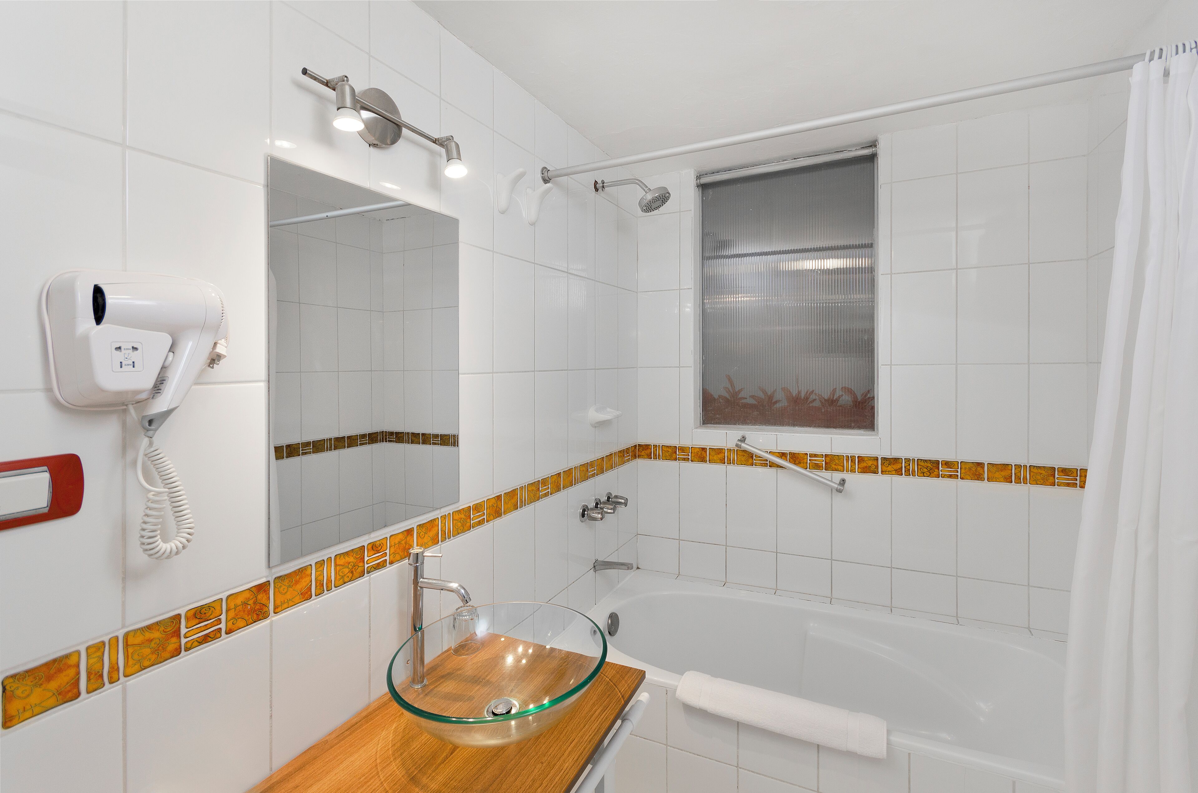 A bathroom with a glass sink, a handheld showerhead, a built-in bathtub, and a hairdryer mounted on the wall.