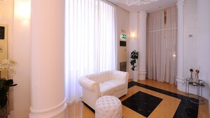 Un sofá blanco, un otoman a juego y una pequeña mesa con palitos de incienso en una habitación luminosa con ventanas grandes y una araña.