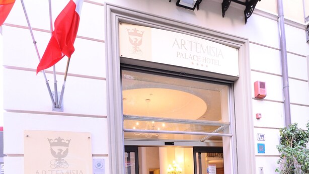 La entrada al Hotel Artemisia Palace, con una puerta de cristal, un cartel y una planta en maceta.