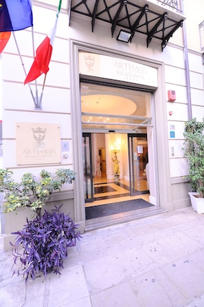 La entrada al Hotel Artemisia Palace, con una puerta de cristal, un cartel y una planta en maceta.