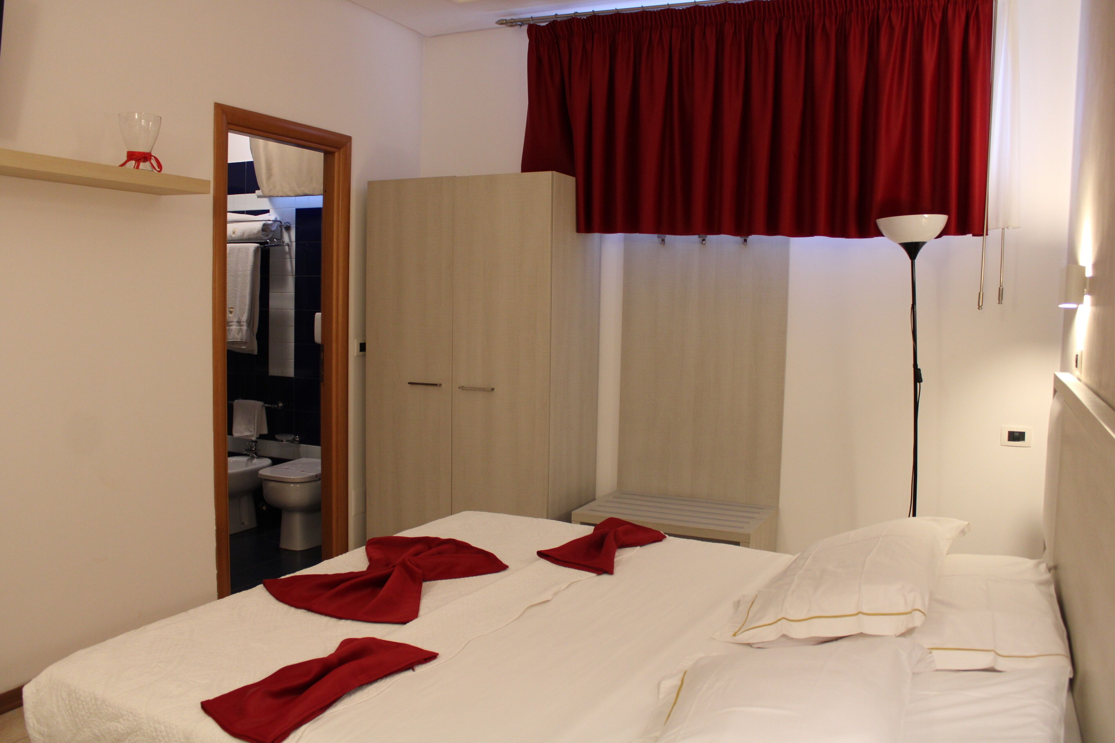 A hotel room with a bed, a wardrobe, a mirror, a red curtain, and a bedside lamp.