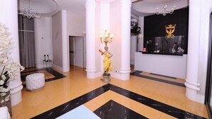 Un vestíbulo con columnas blancas, una estatua, un mostrador de recepción y una araña de cristal.