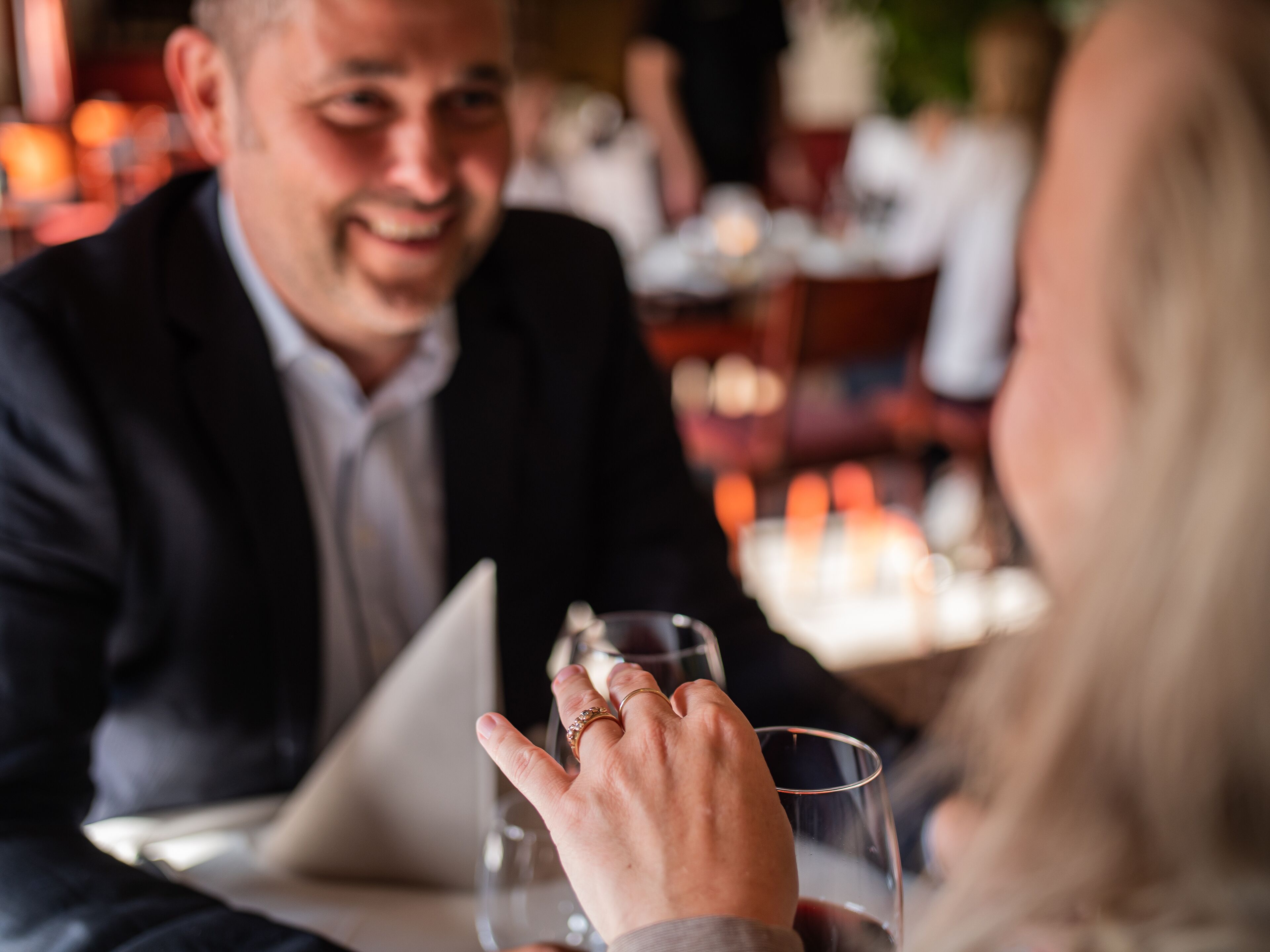 A dining table with a laptop, a glass of red wine, and a person wearing a ring.
