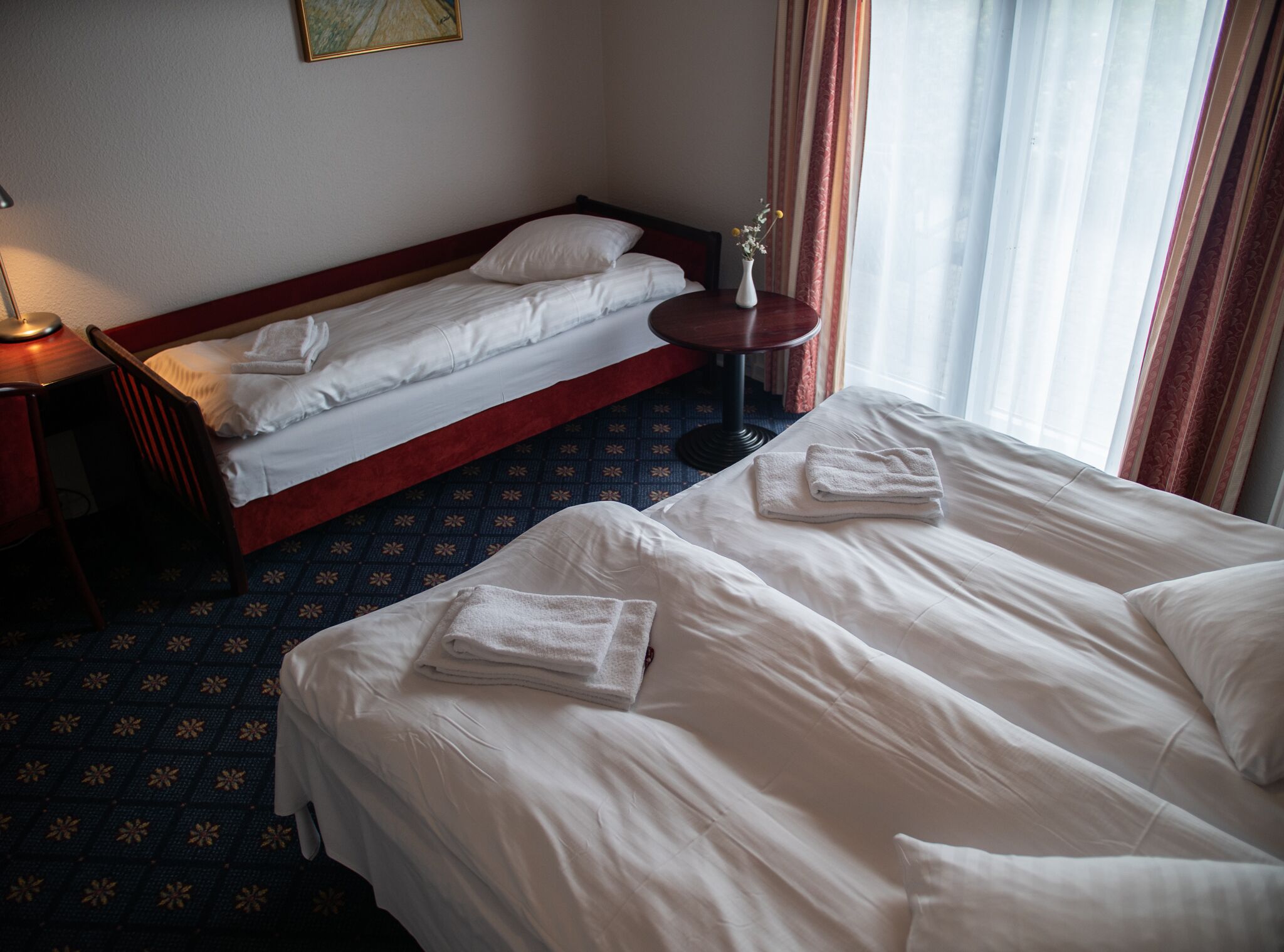 A hotel room with two single beds, a small table with a vase, and a chair.