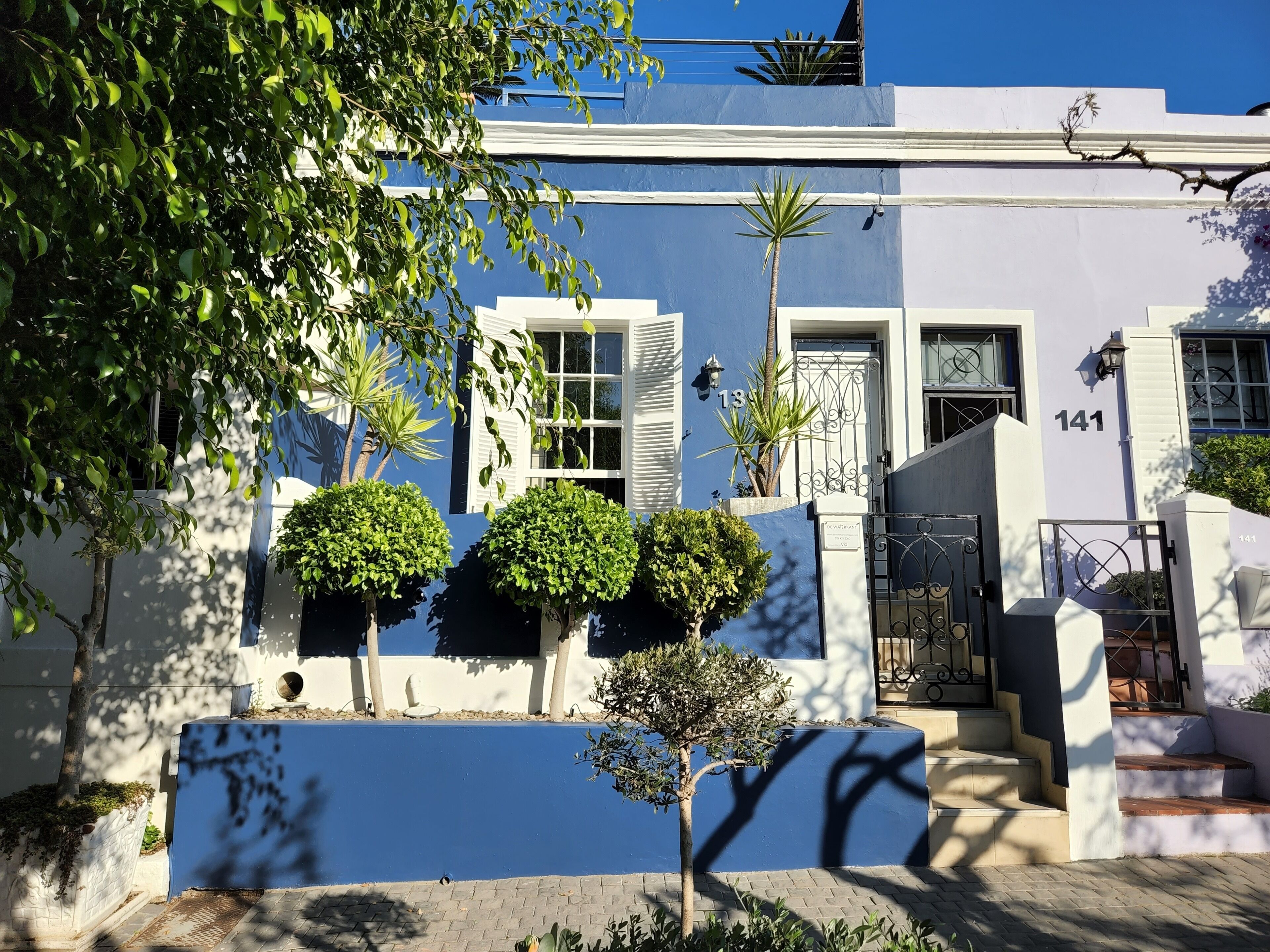 A white building with a blue facade, numbered 141, featuring a gated entrance, a staircase, and a small tree in the foreground.