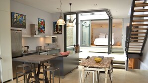 A modern kitchen with a central island, bar stools, and a dining table with chairs. There is a staircase leading to an upper level.