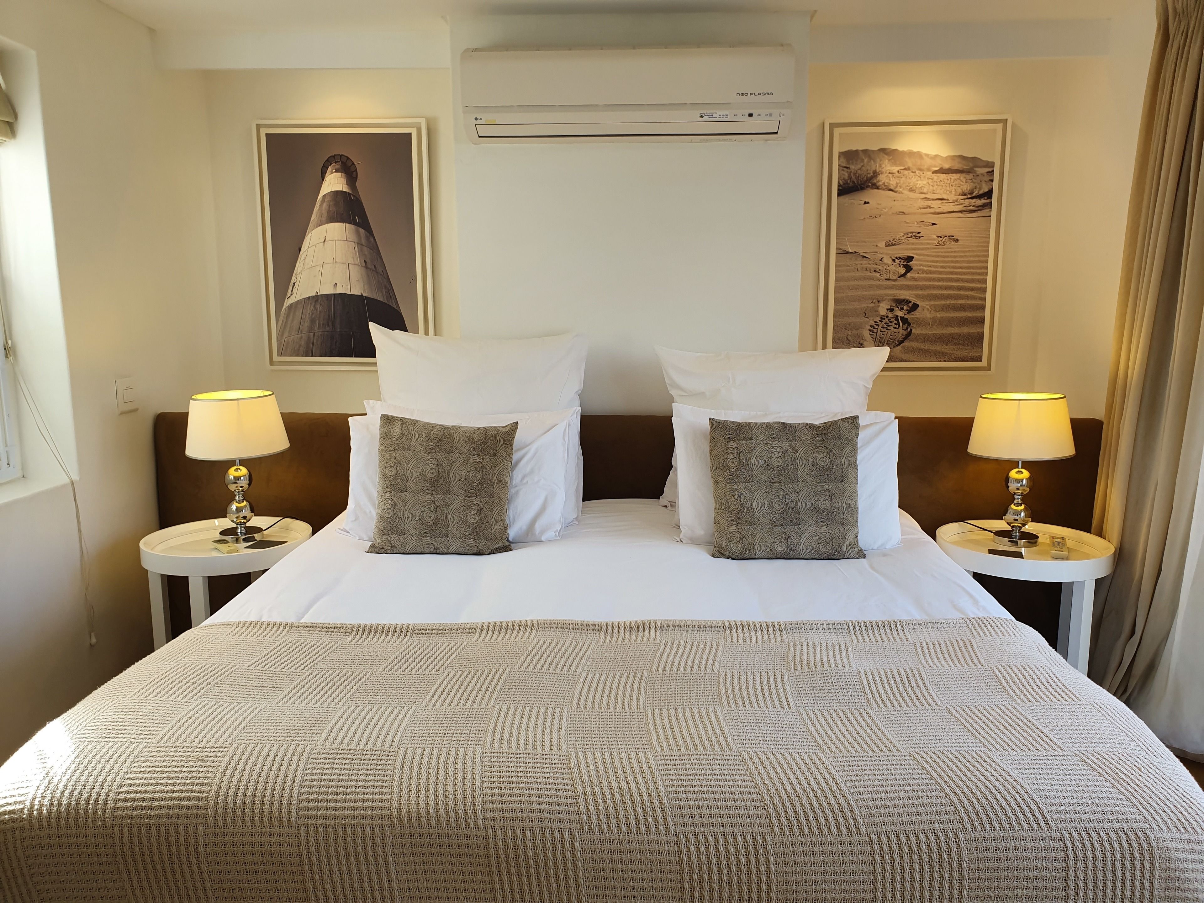 A hotel room with a large bed, two nightstands, a wall-mounted air conditioner, and two framed pictures.
