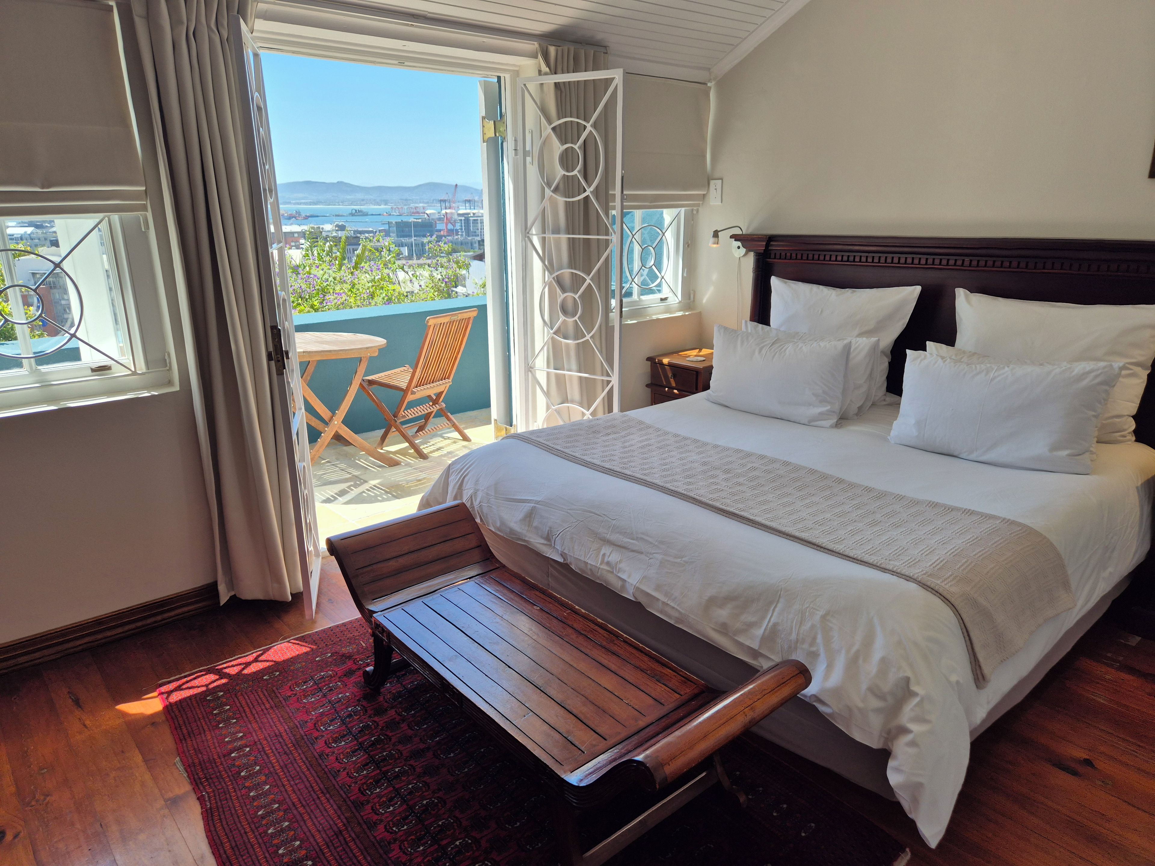 Un dormitorio con una cama grande, un banco de madera, un tapete rojo estampado y vistas a un balcón con una silla.