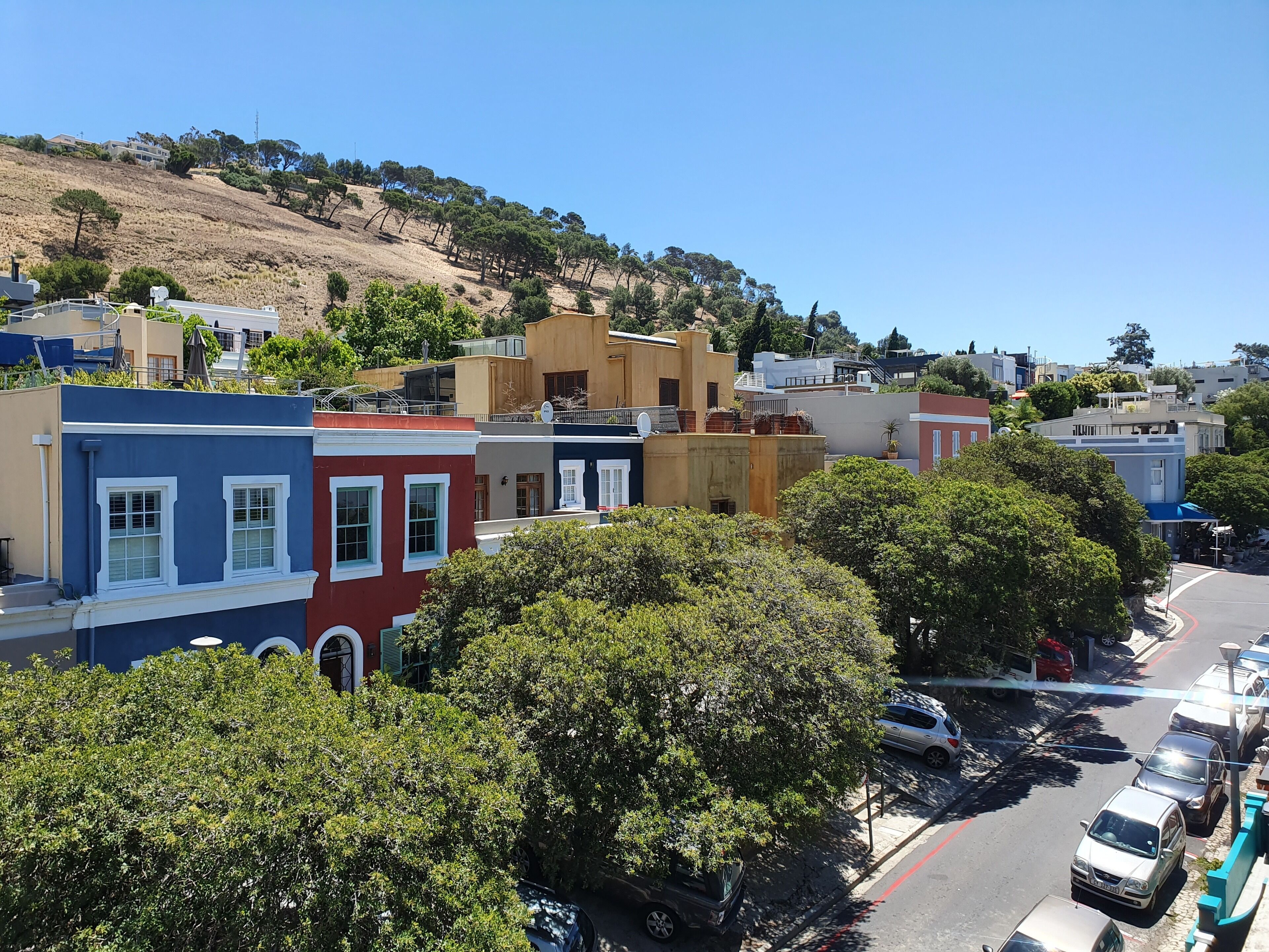 A residential street with colorful houses, parked cars, and a hillside in the background.
