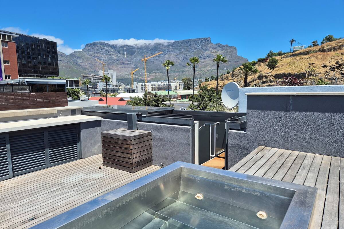 A rooftop with a hot tub, a wooden deck, and a view of mountains and buildings.