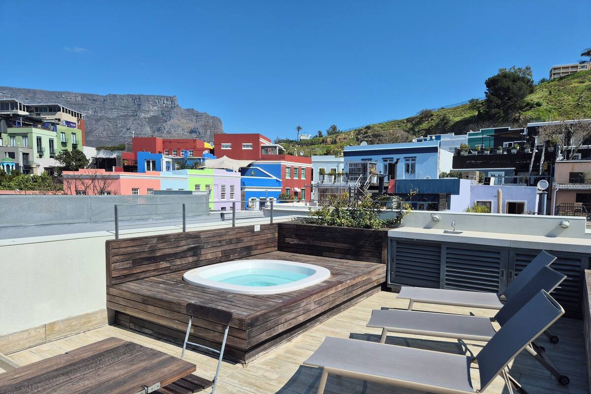 A rooftop terrace with a hot tub, wooden seating, and a view of colorful buildings and a hill.