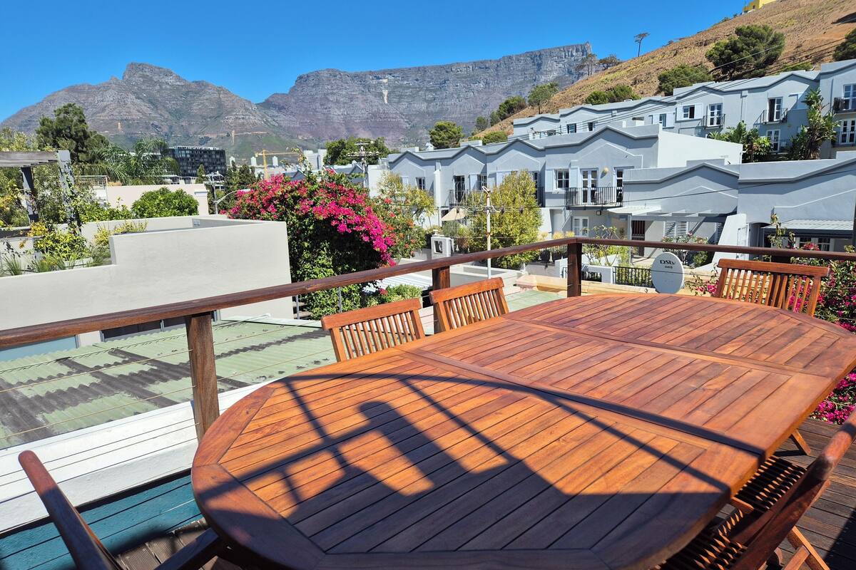 A wooden deck with a table and chairs, overlooking a residential area with houses and a mountain in the background.