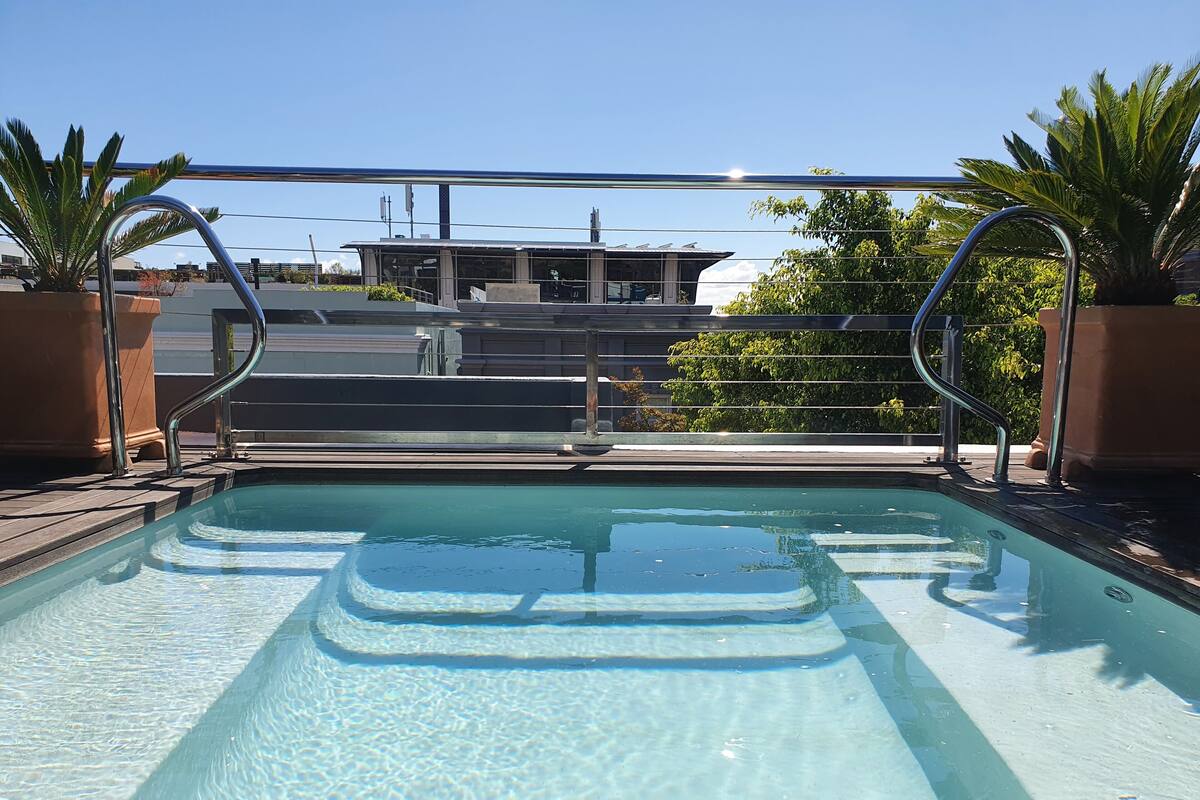 A clear swimming pool with steps leading into it, surrounded by palm trees and a view of buildings in the background.