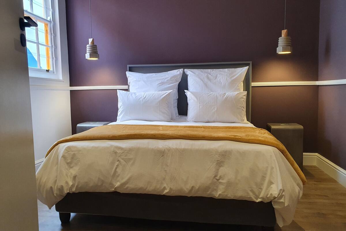 A neatly made bed with white bedding and a brown throw, a nightstand with a lamp, and a window with a view of the sky.