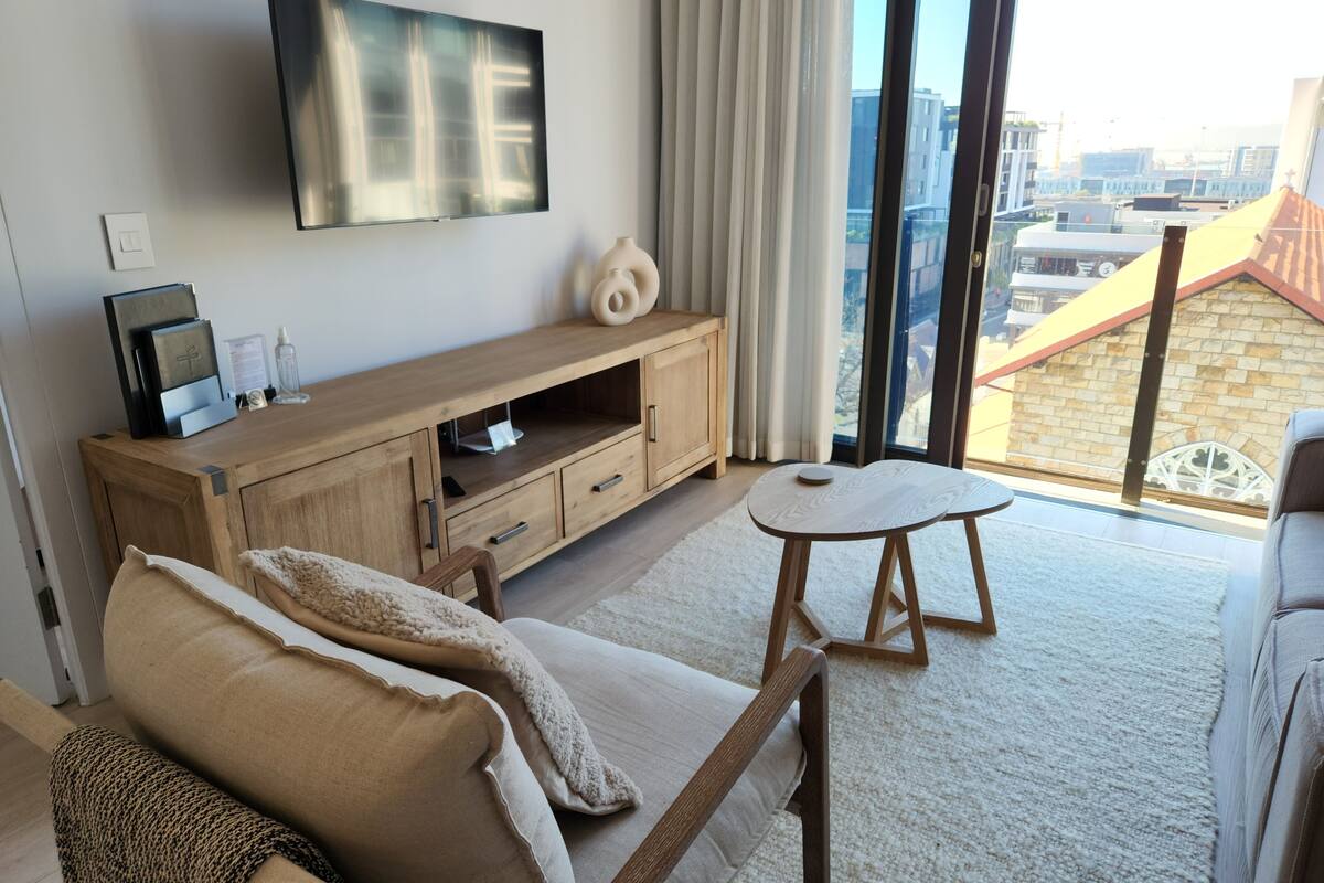 A modern living room with a sofa, a small round table, a wooden TV stand, and a large window offering a city view.