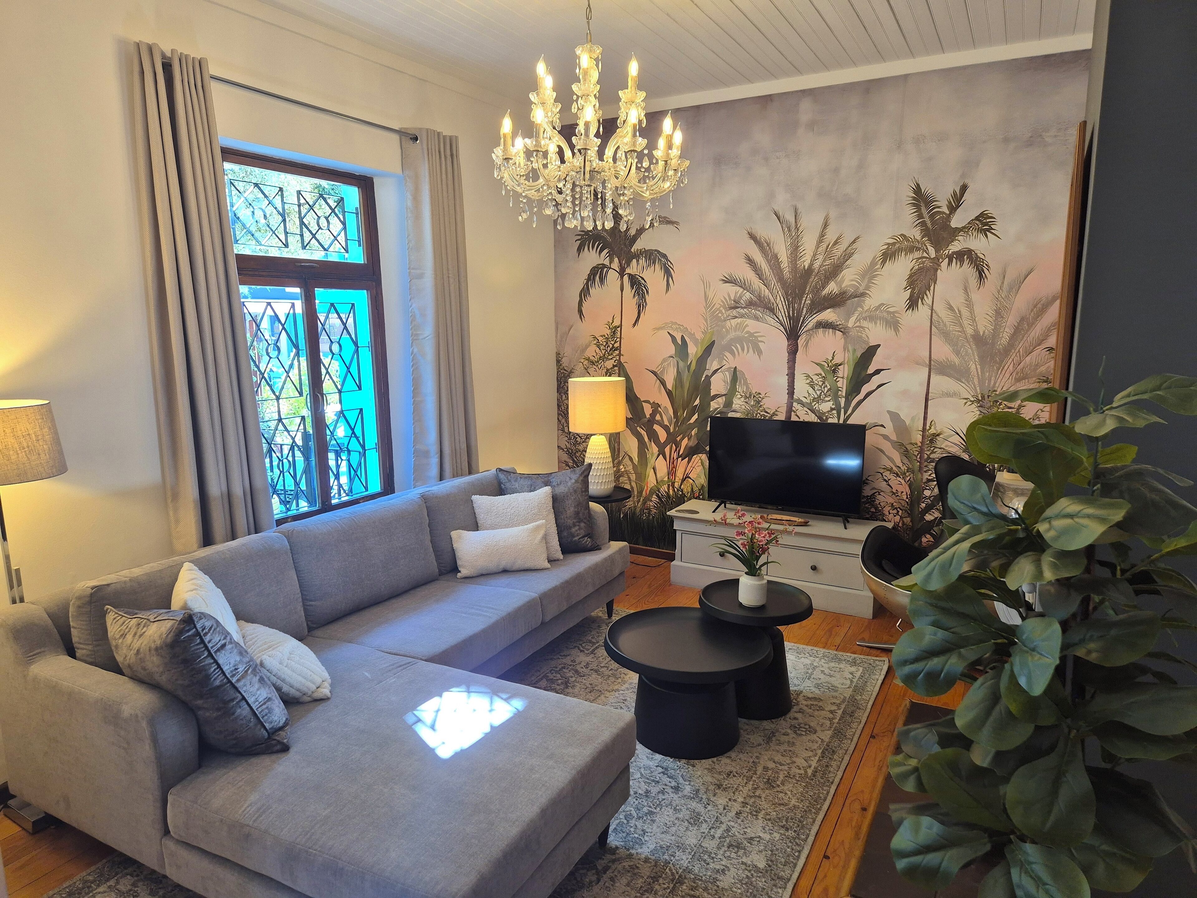 Una sala de estar con un sofá gris, un televisor, una araña y un ventanal con cortinas.