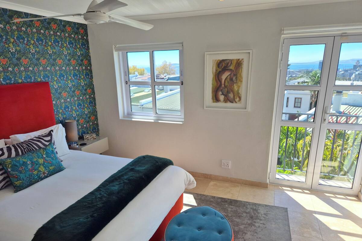 A bedroom with a bed, a red headboard, a blue ottoman, a ceiling fan, a window with a view, and a painting on the wall.