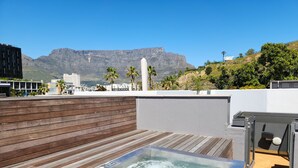 A rooftop with a hot tub, wooden deck, and a view of mountains and buildings.